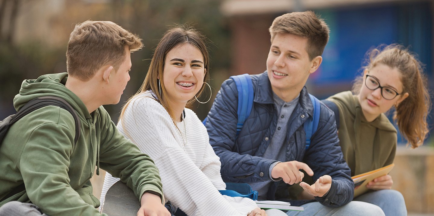 Eine jugendliche Schülergruppe (zwei Jungen, zwei Mädchen) sitzen auf einer Bank oder einer Mauer und unterhalten sich. Alle haben dunkelblondes bis brünnettes Harr. Die JUngen haben einen Kurzhaarschnitt die Mädchen langes Haar. Das Mädchen ganz rechts trägt zudem Brille und einen Zopf