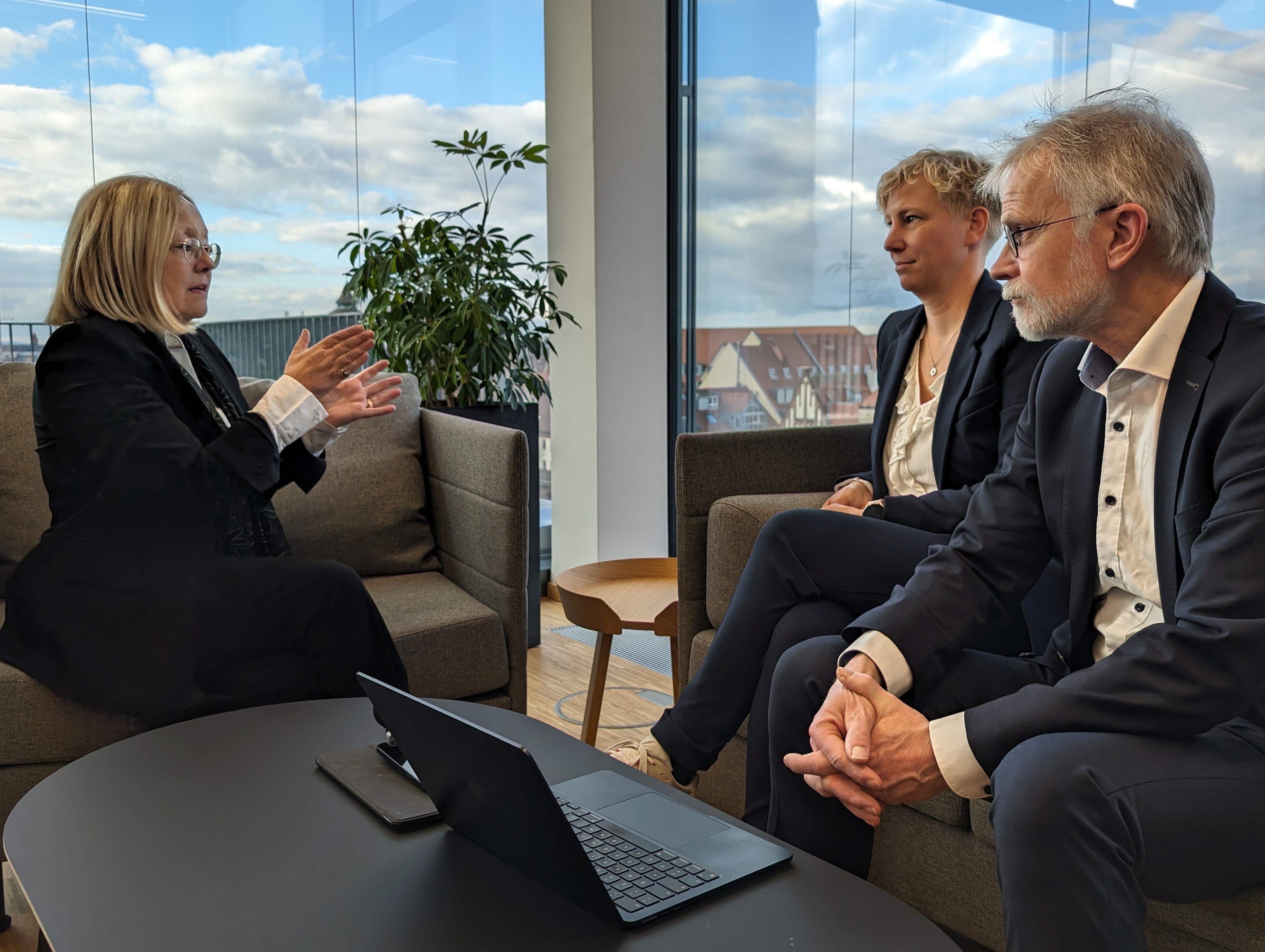 Dr. Irmgard Stippler in dunklem Anzug gibt Redakteuren von G+G in dunklen Anzügen ein Interview. Sie sitzen an einem Fenster. Frau Stippler gestikuliert