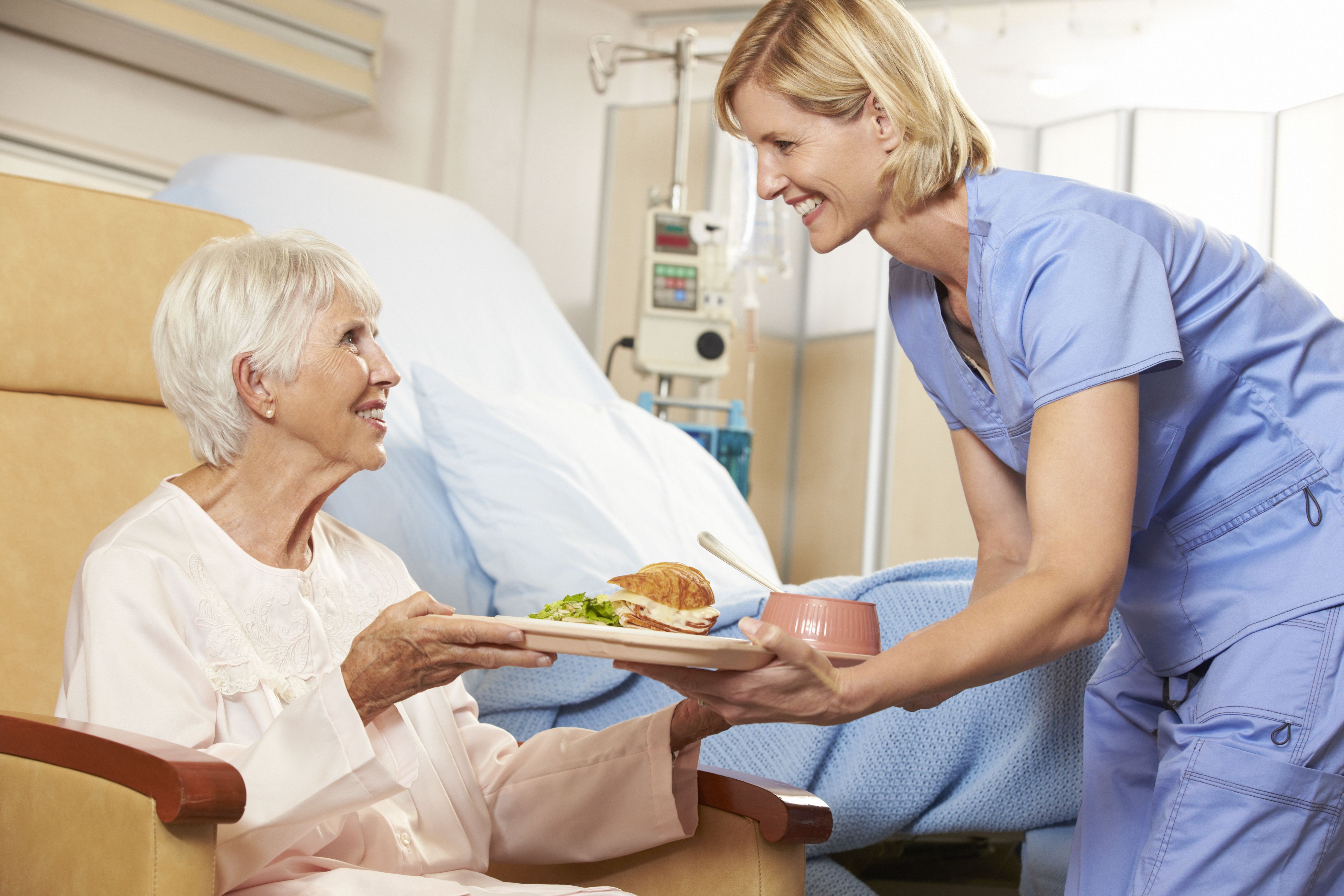 Schmuckfoto zeigt eine ältere Frau im Krankenhausbett sitzend, der ein Tablett mit Essen von einer Fachkraft gereicht wird.