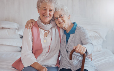 Foto: Zwei ältere Frauen sitzen auf einem Bett und umarmen sich.