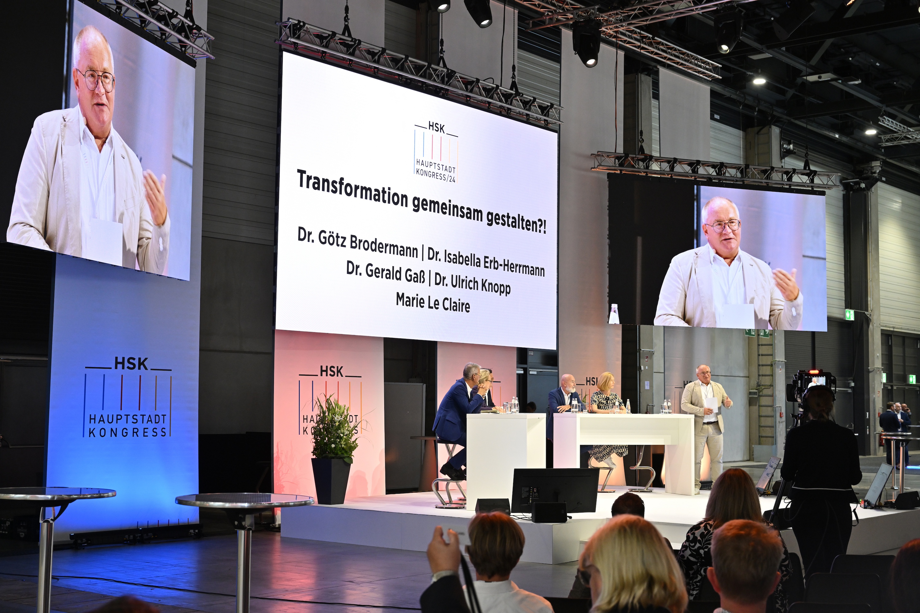 Das Bild zeigt die Totale einer Bühnen mit drei großen videoleinwänden. Darunter bedindet sich eine Theke mit Podiumsteilnehmern, rechts danebenn ein Redner-Pult