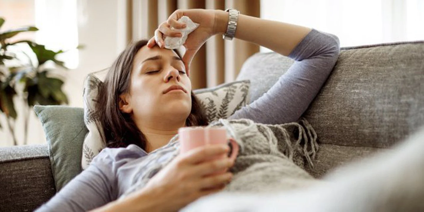 Das Bild zeigt eine Junge Frau, die offenbar krank und erschöpft auf einer grau gemusterten Couch liegt. In der rechten Hand hält sie eine pinkfarbene Teetasse. Ihre linke Hand hält ein Taschentuch und liegt an der Stirn. Im Hintergrund ist ein Fenster mit Zimmerpflanzen zu sehen.