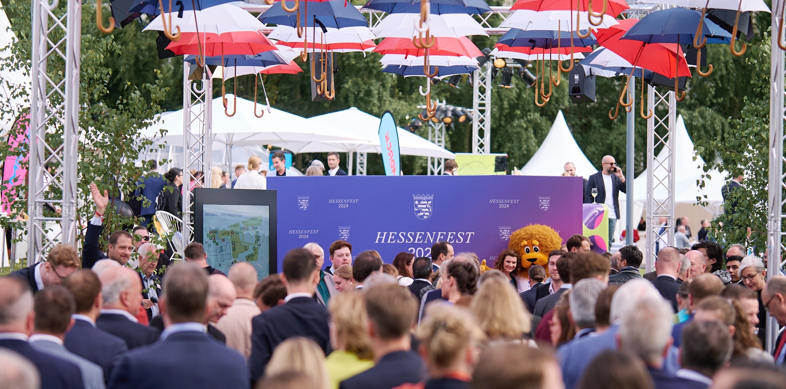 Das Bild zeigt eine große Menschenmenge auf dem Hessengfest. Oben sind rote, weiße und blaue Regenschirme zur Dekoration aufgehängt. ImHintergrund sieht man große weiße Sonnenschirme und weiße Festzelte.