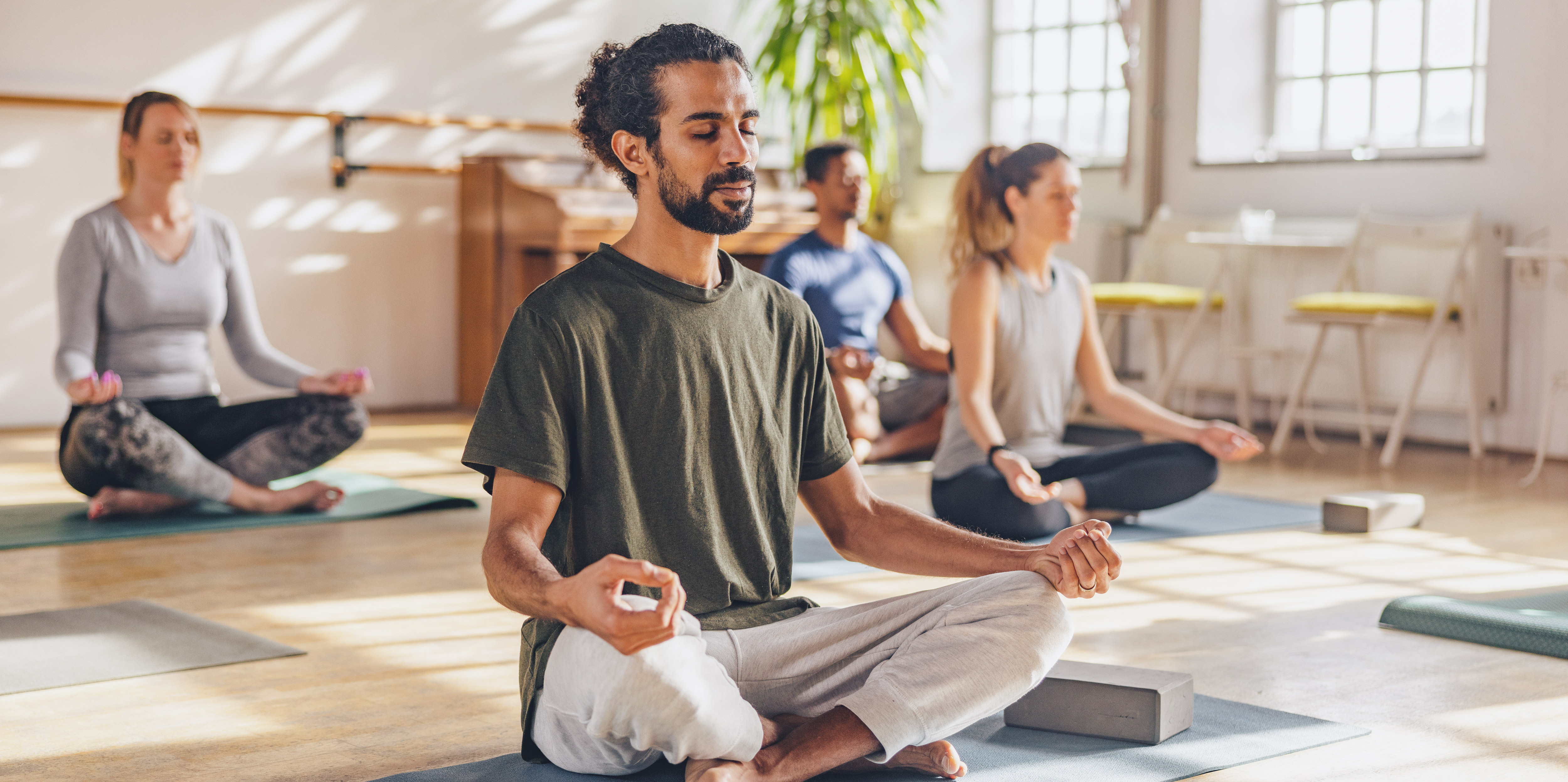 Das Bild zeigt eine vierköpfige Yoga-Gruppe im Lotussitz. Im Vordergrung ein Mann mit dunklen Haaren, Dutt und Vollbart, im Hintergrund zwei FRauen und ein weiterer Mann.