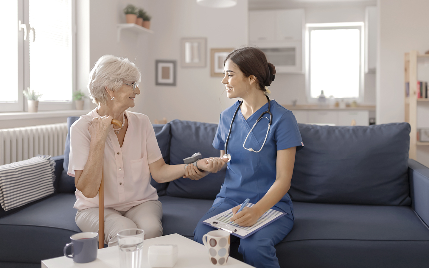 Foto zeigt eine Patientin (links) mit einer Pflegerin (rechts) im Gespräch. Beide sitzen auf einem Sofa in einer hellen Wohnung. Die Pflegerin trägt blaue Berufskleidung.