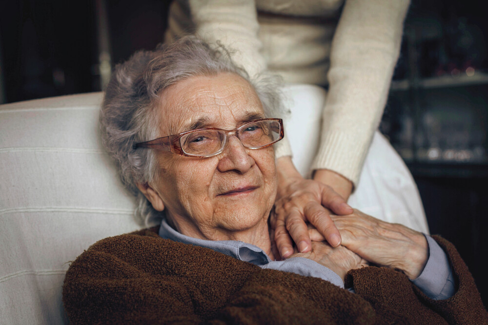 Eine alte Frau auf einer Liege, aus dem Hintergrund hält jemand ihre Hand 