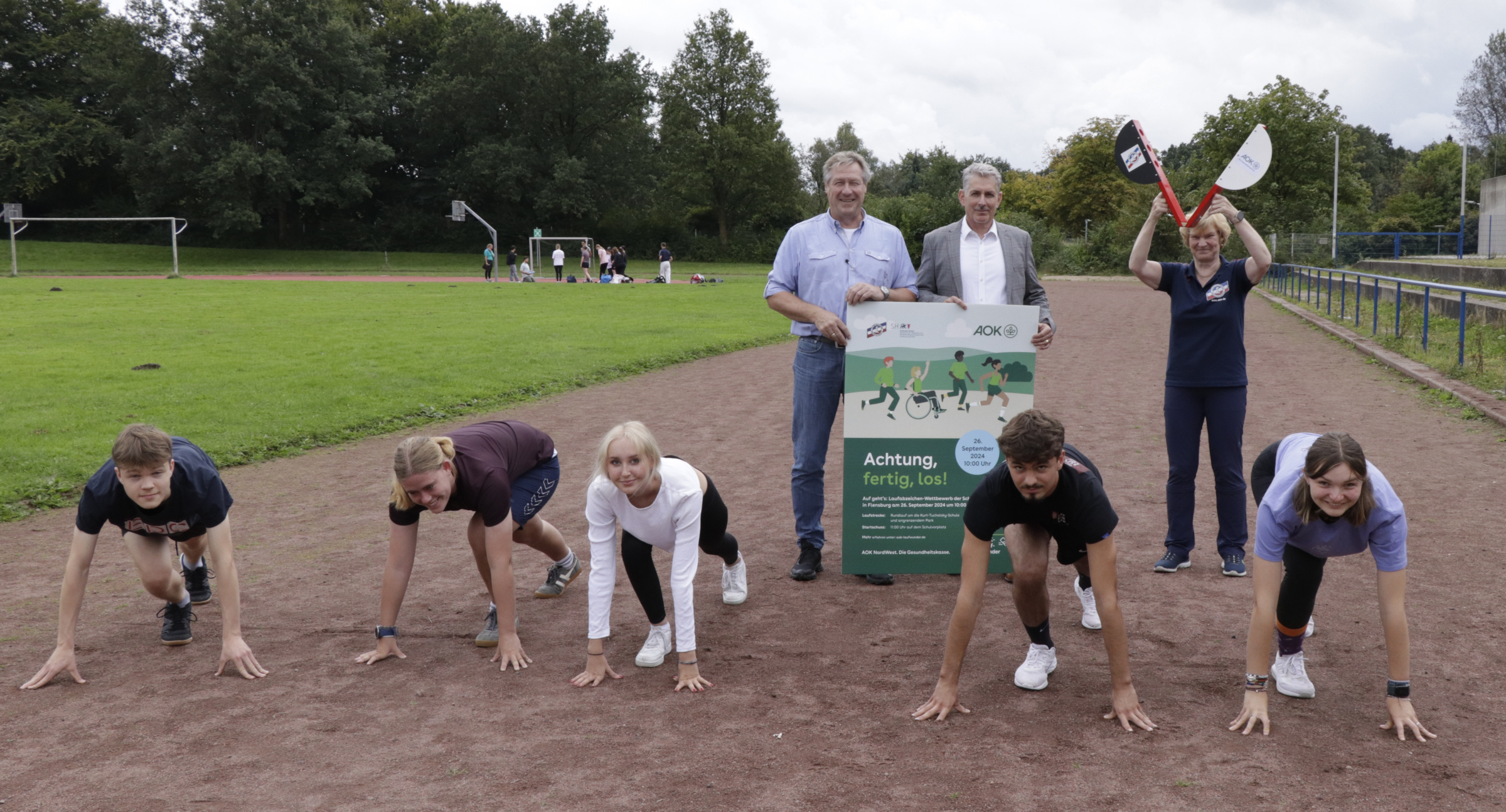 Foto zeigt einen Schul-Sportplatz, auf der Aschebahn haben sich fünf Schülerinnen und Schüler in Startposition gebracht, dahinter stehen Schulleiter und AOK-Repräsentant mit dem Plakat zum Lauftag sowie rechts danaben die Vertreterin des SHLV mit einer Startklappe.