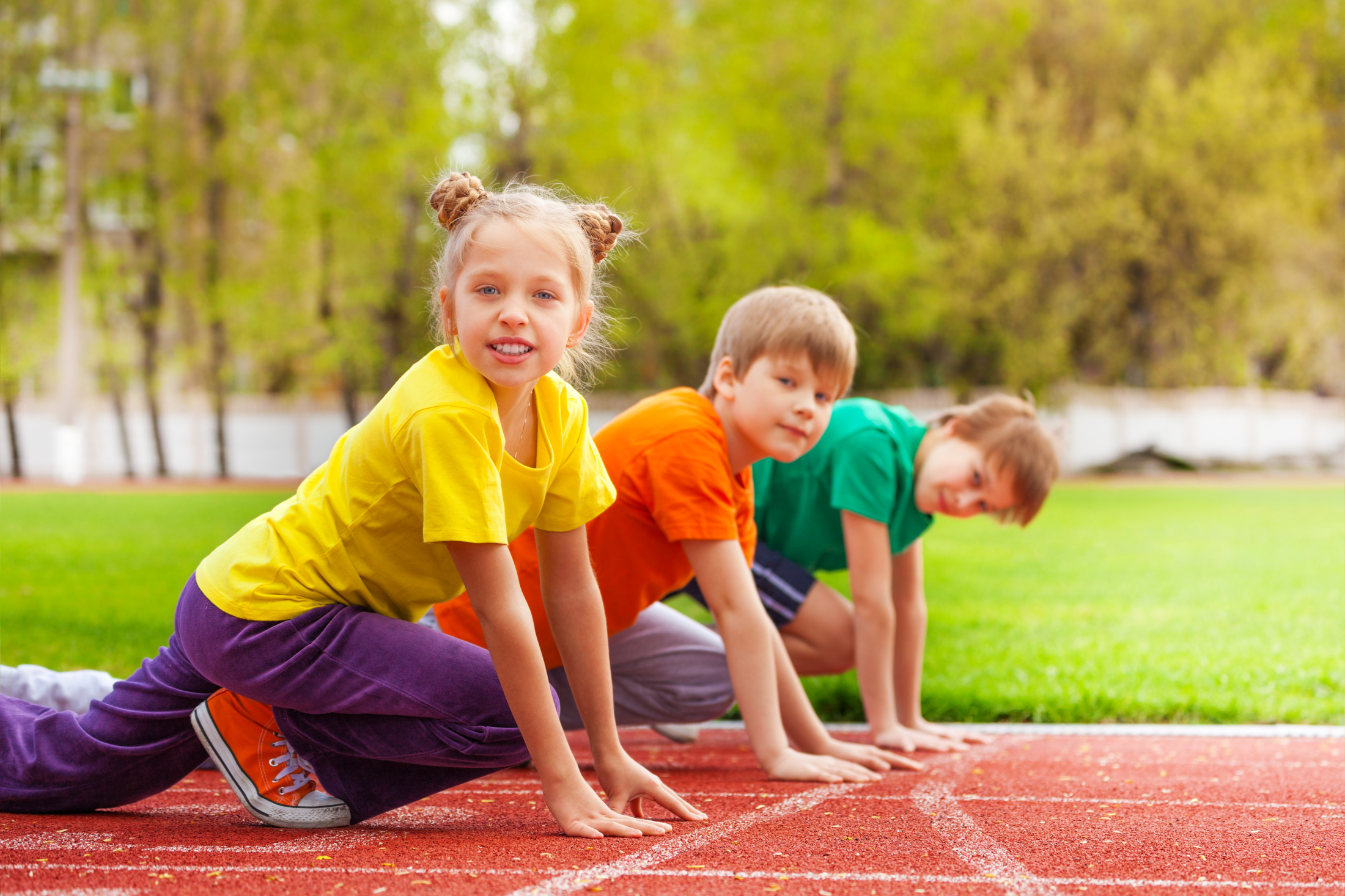 Das Foto zeigt ein (Schul-)Sportgelände; im Vordergrund sind eine Schülerin und zwei Schüler zu sehen, die auf einer Tartanbahn die Startposition zum Loslaufen eingenommen haben und lächelnd in die Kamera schauen.