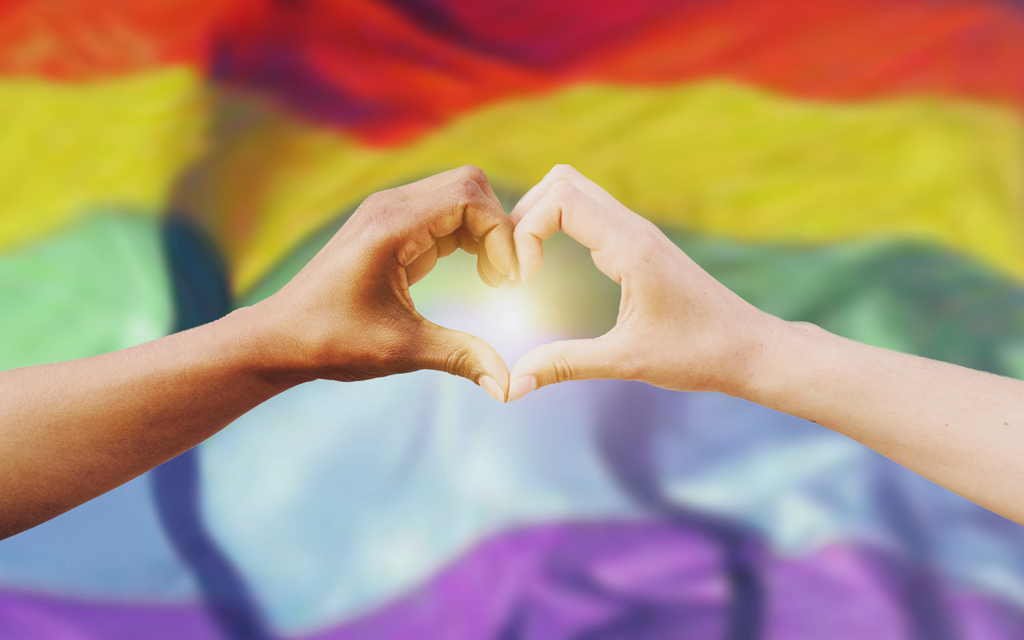 Vor dem Hintergrund einer Regenbogenfarbe sieht man eine farbige Hand und eine helle Hand, die zusammen eine Herzform bilden.
