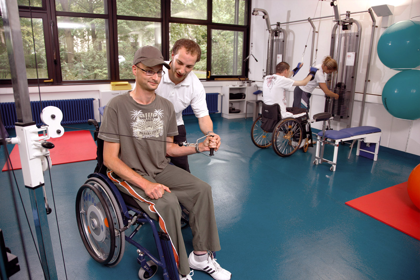 Foto: Patienten in Rollstühlen trainieren in einem Trainingszentrum. Im Vordergrund unterstützt ein Physiotherapeut bei einer Übung.