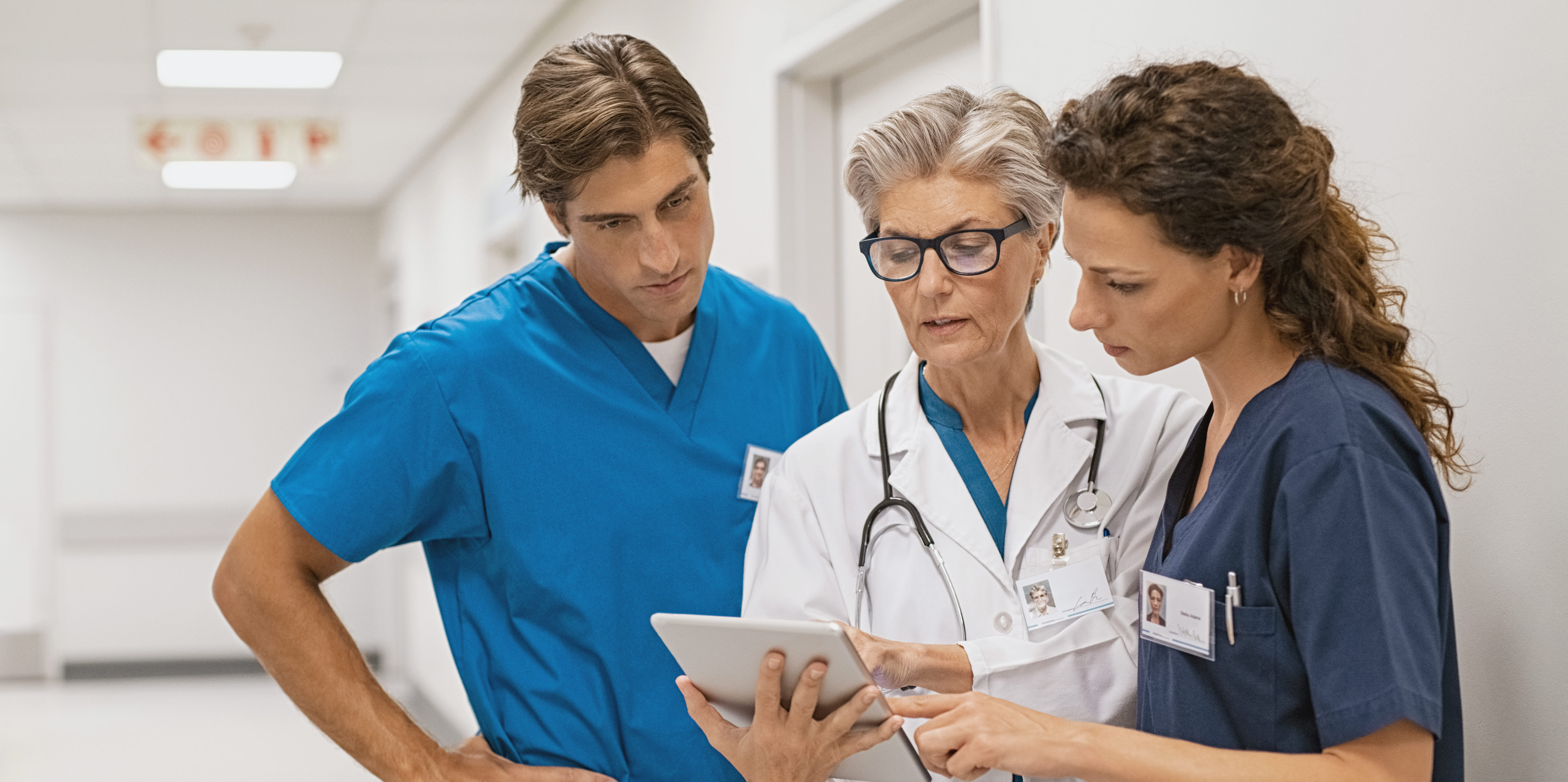 Das Bild zeigt eine Ärztin (Mitte mit weißem Kittel und Stethoskop um die Schulter) einen Pfleger (links) und eine Pflegerin (rechts) bei einer Teambesprechung auf dem Klinikgang. Die Pflegefachpersonen tragen grünblaue Arbeitskleidung.