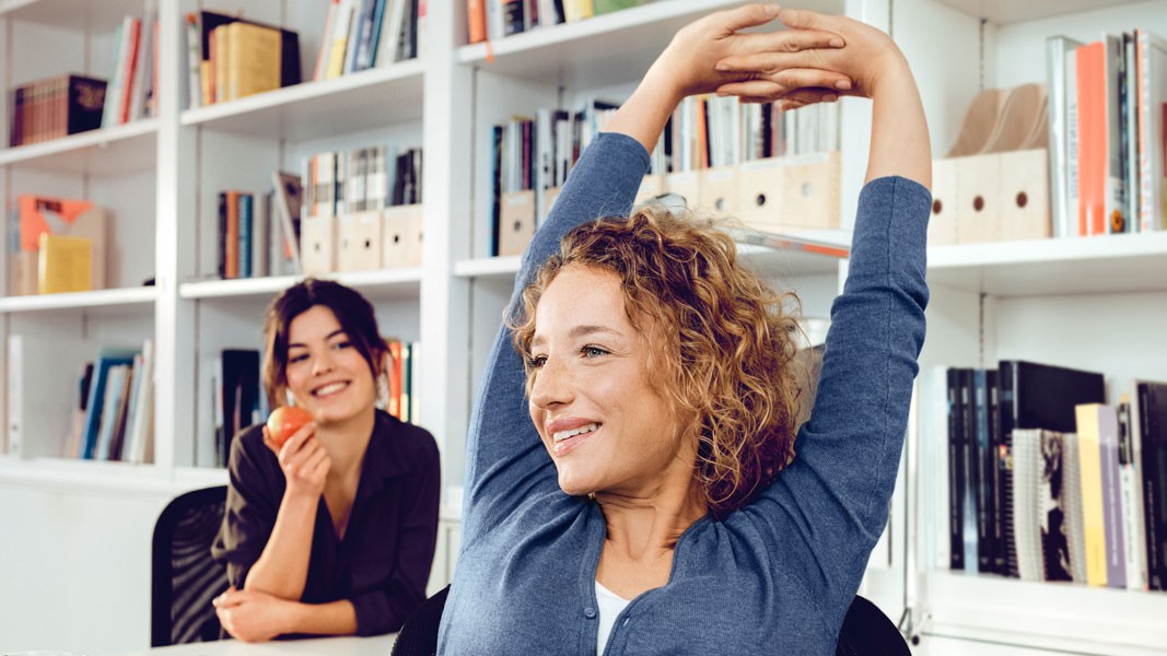 Eine Frau sitzt auf einem Bürostuhl und streckt sich. Im Hintergrund sitzt eine weitere Frau und hält einen Apfel in der Hand.