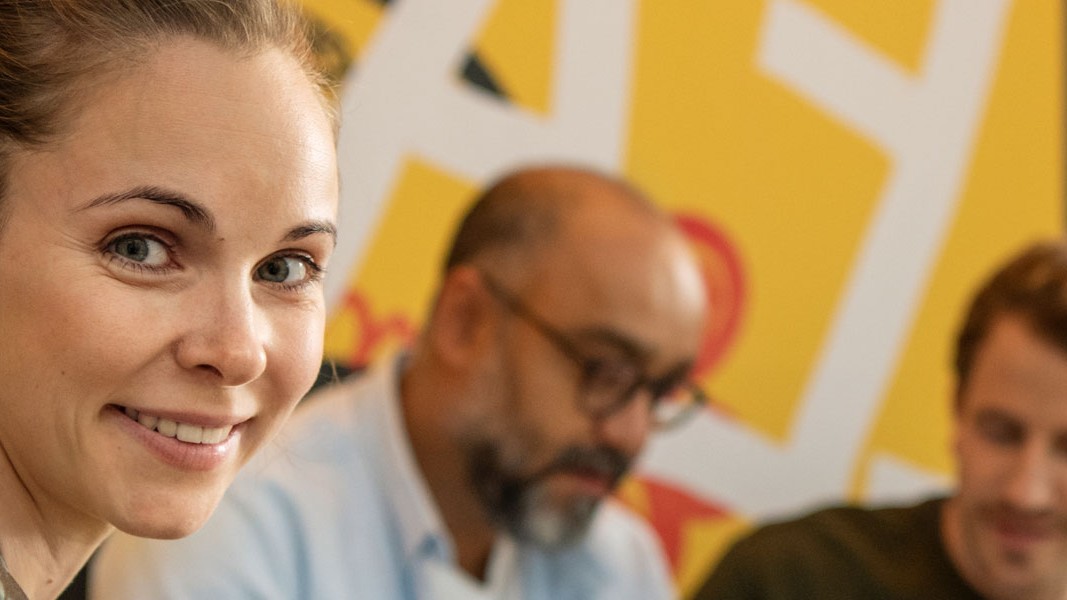 Eine Frau lächelt im Vordergrund in die Kamera. Zwei Männer sitzen im Hintergrund.