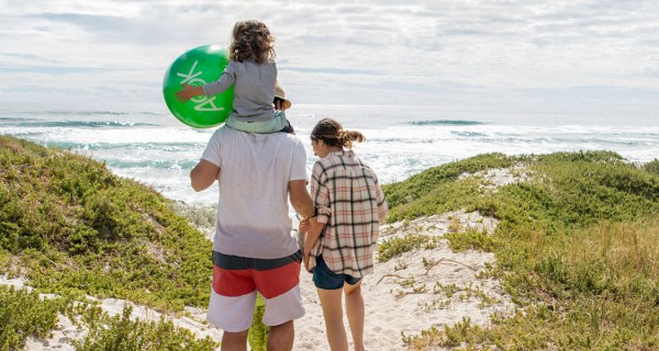 Mann und Frau stehen vor Dünen, ein Kind sitzt auf den Schultern des Mannes und hält einen Ball. Im Hintergrund ist das Meer.