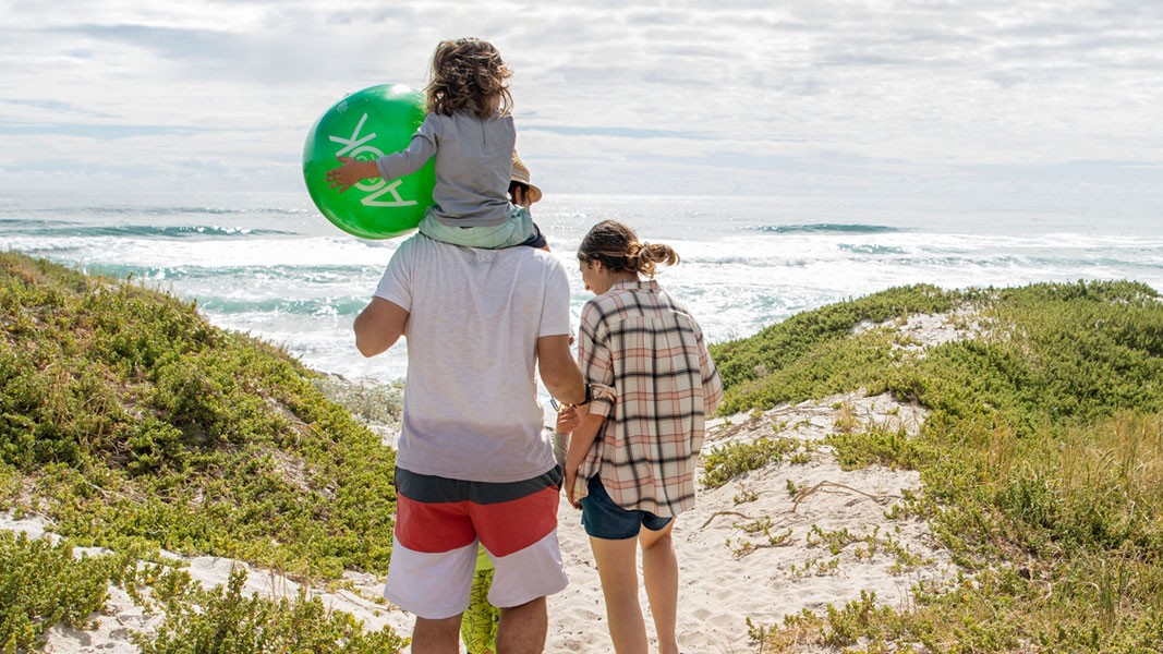 Mann und Frau stehen vor Dünen, ein Kind sitzt auf den Schultern des Mannes und hält einen Ball. Im Hintergrund ist das Meer.